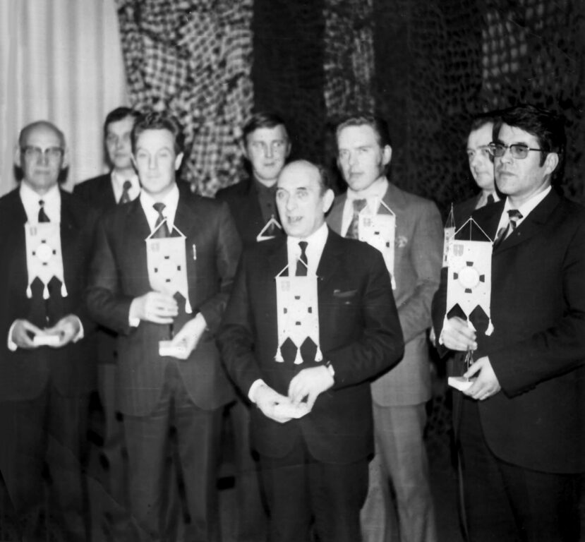 Kahdeksan henkilöä juhla-asussa pitelemässä pöytästandaareja Kauhavan Reservinaliupseerien vuosikokouksessa 1973.